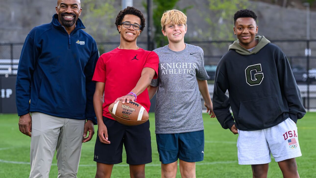 A coach stands with three boys on a football field. One boy holds a football. They are dressed in casual sportswear.
