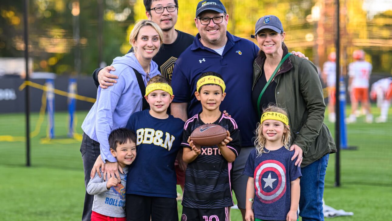 A group of adults and children stand on a grass field. The children wear yellow headbands, and one is holding a football. Players practice in the background.
