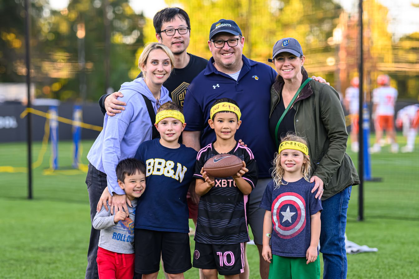 A group of adults and children stand on a grass field. The children wear yellow headbands, and one is holding a football. Players practice in the background.