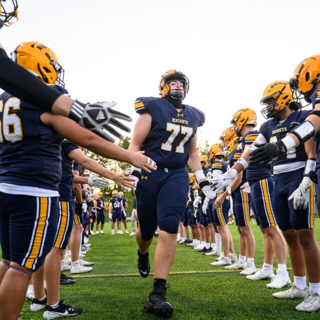 Football player in a number 77 uniform runs through a line of teammates who are clapping and extending their hands for high-fives, on a grassy field.