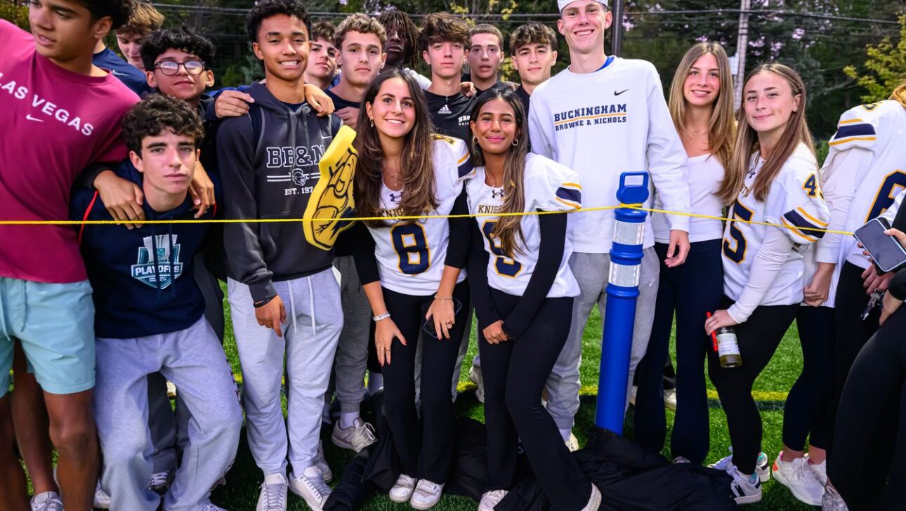 A group of people, some in sports jerseys, stand together on a sports field. A yellow rope is in front of them. Trees are visible in the background.