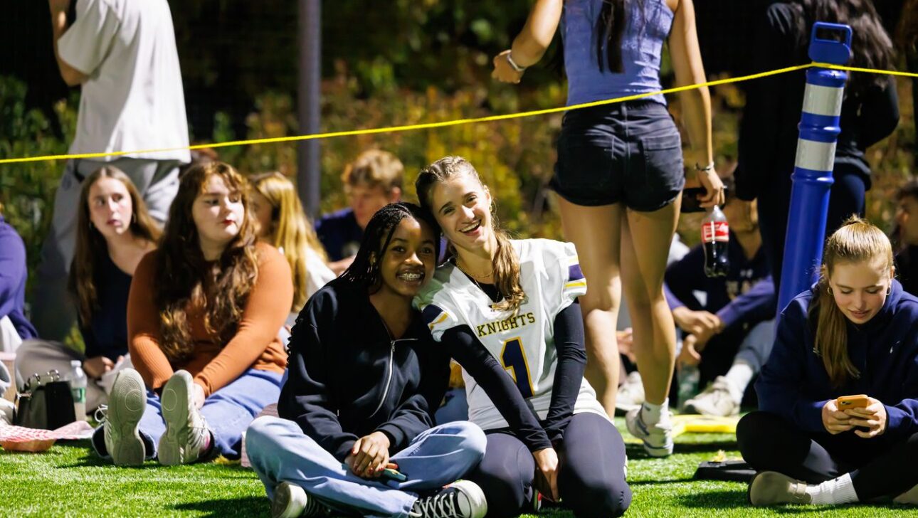 A group of young people sit on grass at an outdoor event. Two of them smile at the camera, with one wearing a "Knights" jersey.