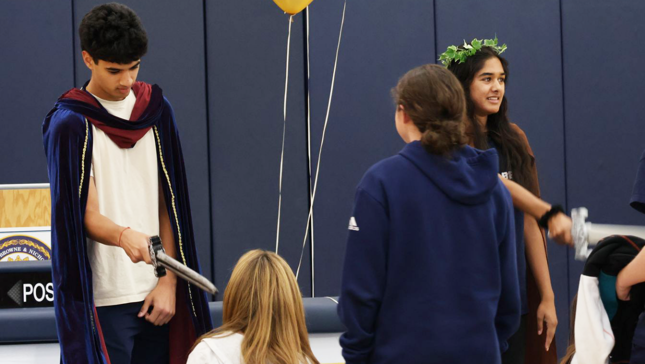 A group of young people are dressed in costumes performing a play. One holds a sword, and another wears a leafy crown. Balloons are in the background.