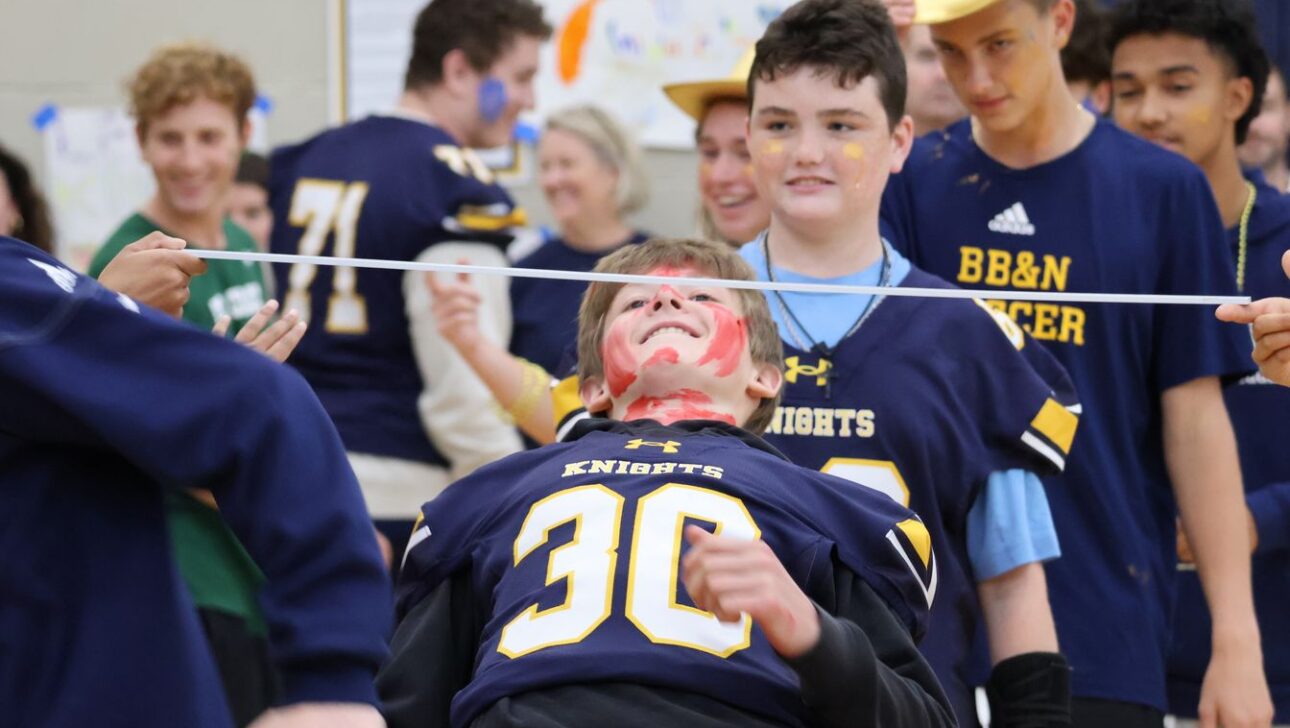 Students participate in a limbo game, with one student bending backwards under a horizontal bar. Others watch and cheer. They're wearing jerseys and celebratory face paint.