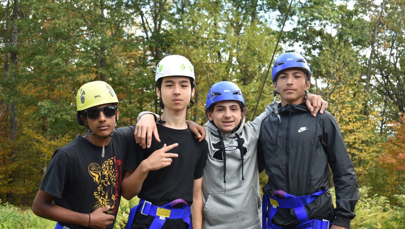 Four teenagers wearing helmets and harnesses stand together outdoors with trees in the background, posing for the camera.