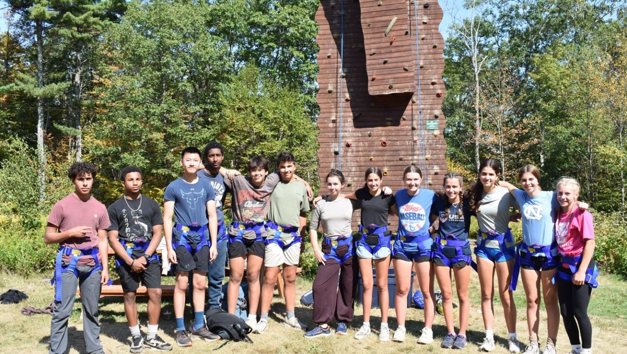 A group of teenagers wearing harnesses stand in front of a wooden climbing wall outdoors, surrounded by trees on a sunny day.