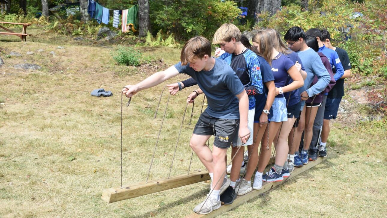 A group of teenagers stand on two long wooden planks, holding ropes for balance, participating in an outdoor team-building activity on grass.