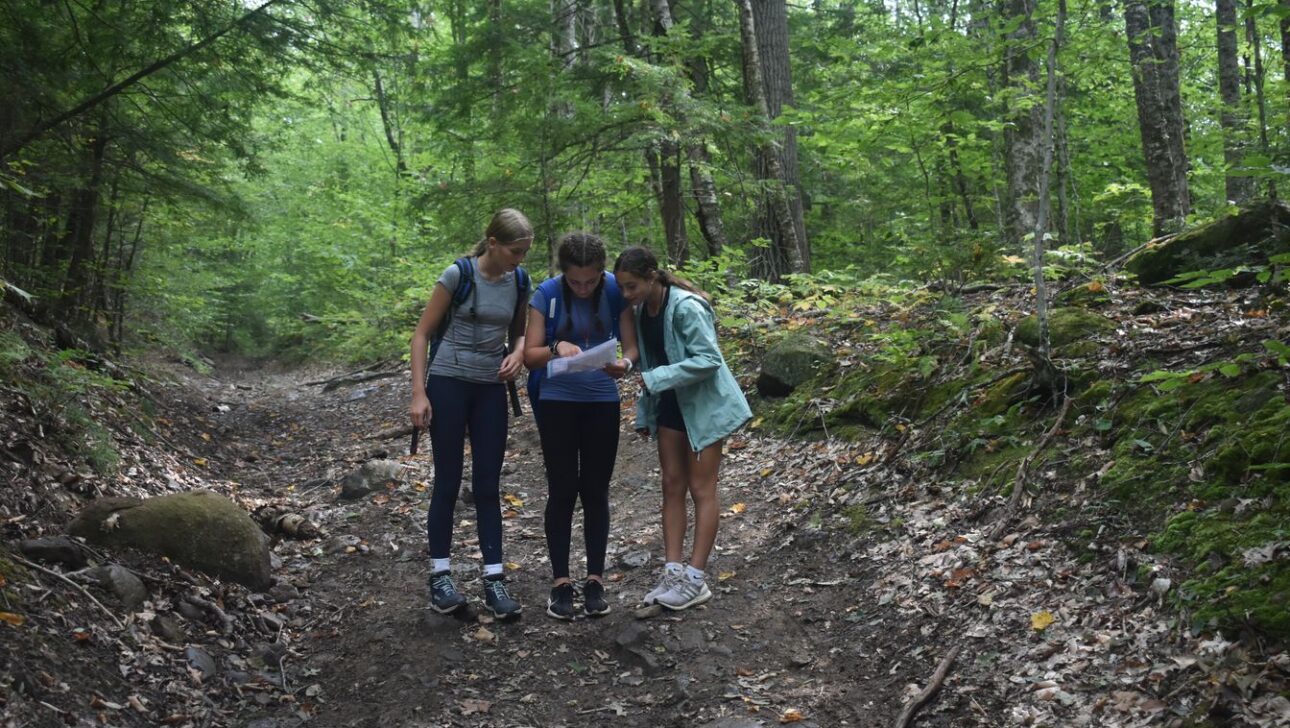 Three girls stand on a forest trail looking at a map, surrounded by trees and greenery.