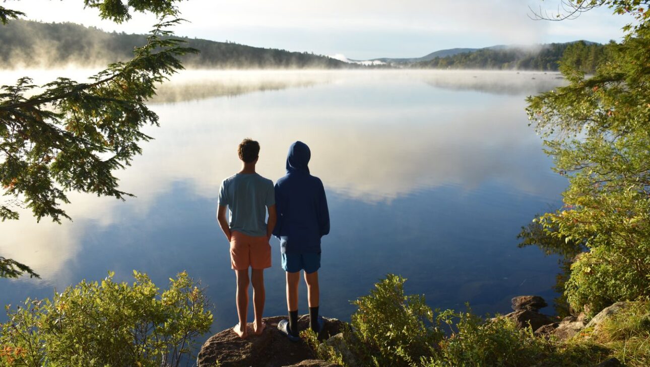 Two people stand on rocks at the edge of a calm lake, facing the water, surrounded by greenery with mist rising in the distance.