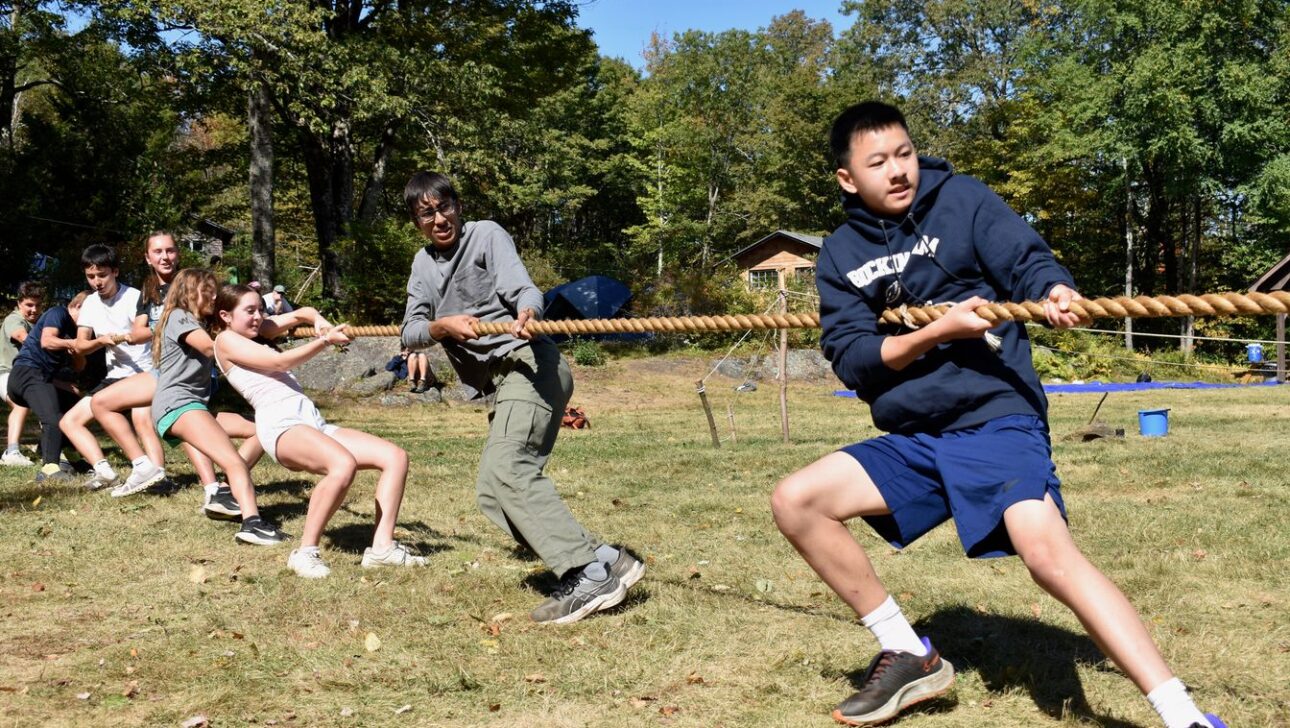 A group of people outdoors participate in a tug-of-war game, pulling on a thick rope, with trees and small buildings in the background.