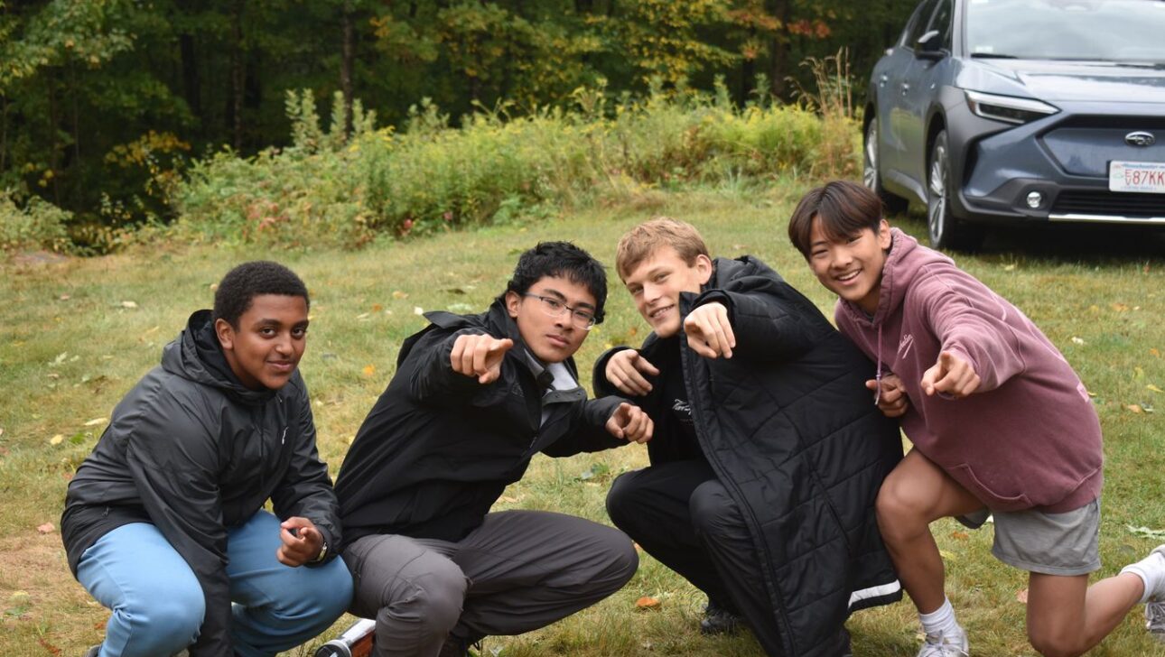 Four young people kneel on grass outdoors, pointing at the camera, with trees and a parked car in the background.