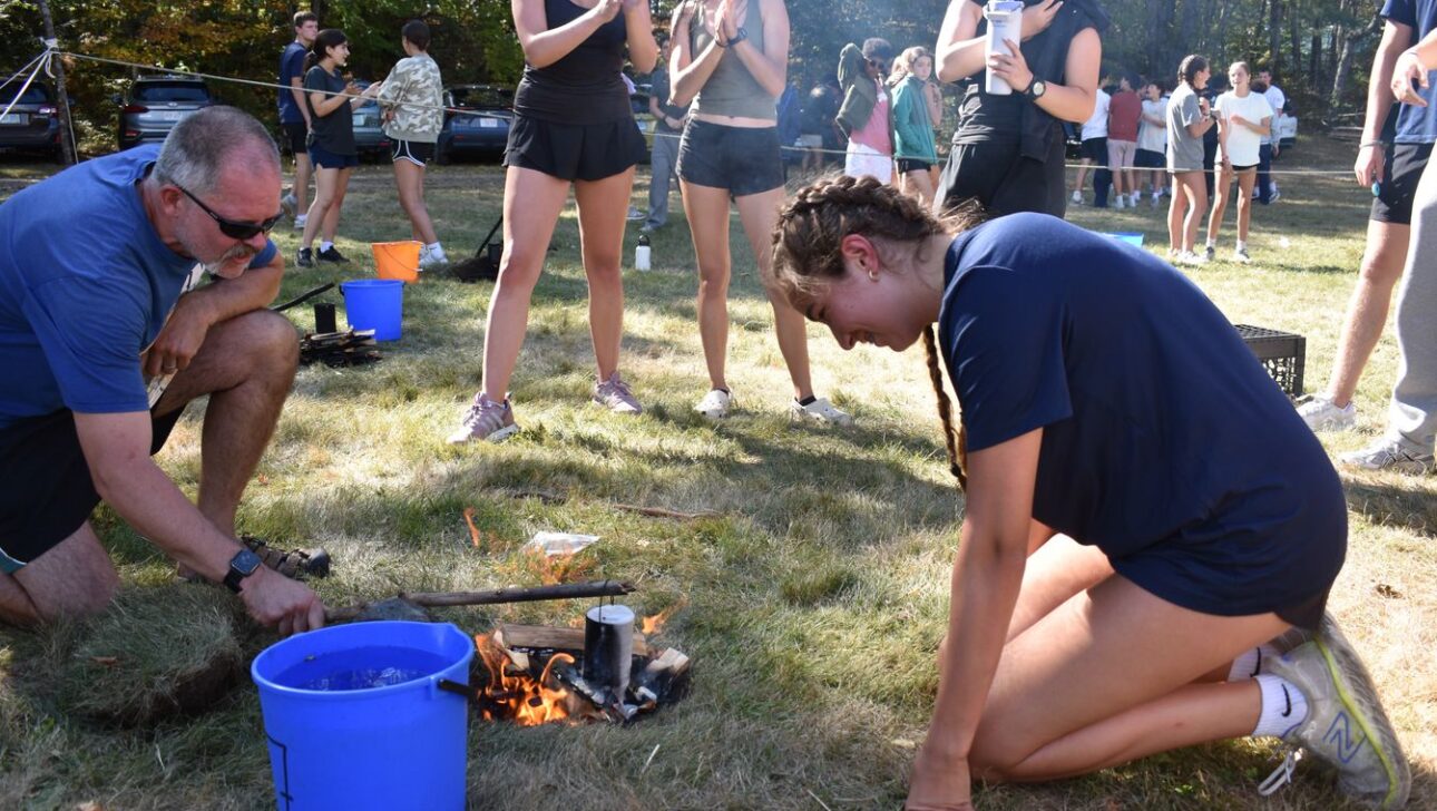 A man and a young woman kneel by a small campfire outdoors, with a blue bucket nearby. Several people stand in the background, some clapping and others watching.