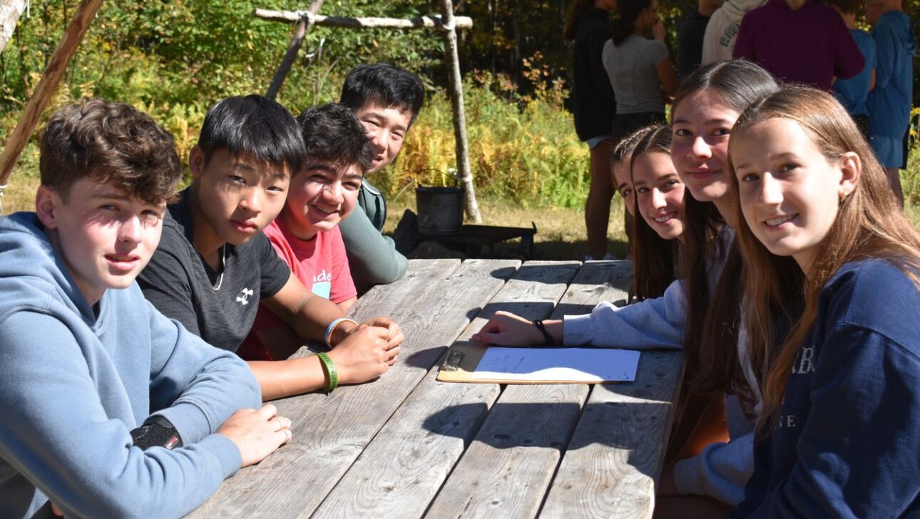 A group of teens sit around a wooden picnic table outdoors on a sunny day, with papers and a pen in front of them. Other people are seen standing in the background.