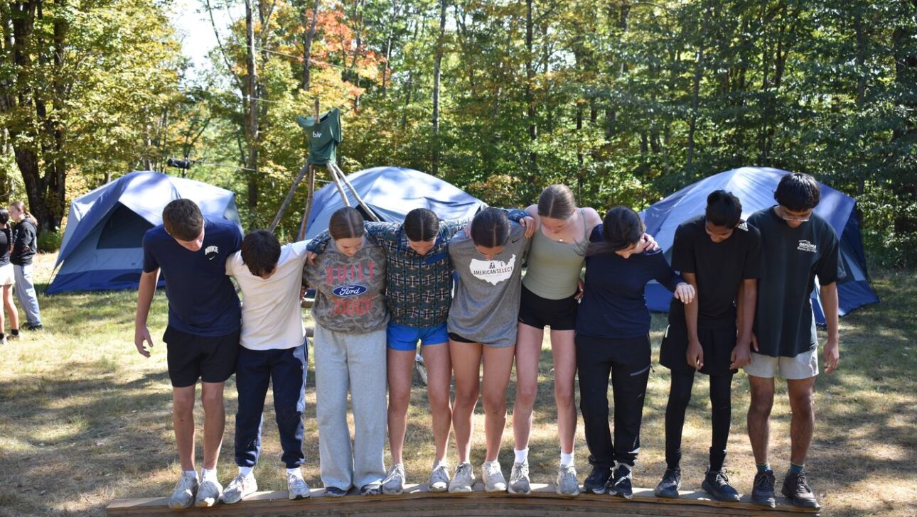 A group of eight people stand arm-in-arm on two wooden planks in a grassy area with tents and trees in the background, participating in an outdoor activity.