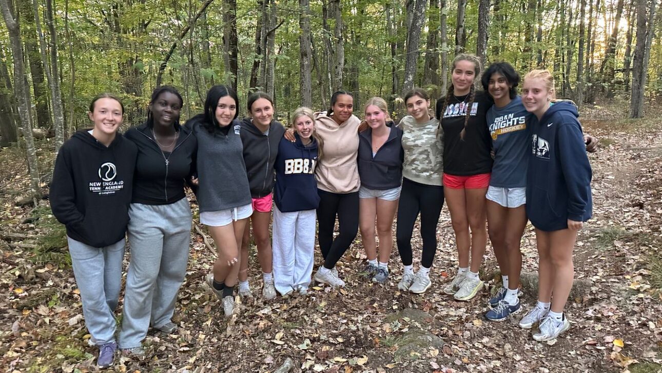 A group of eleven young people stands together outdoors in a forested area, smiling at the camera. They are dressed in casual athletic clothing.