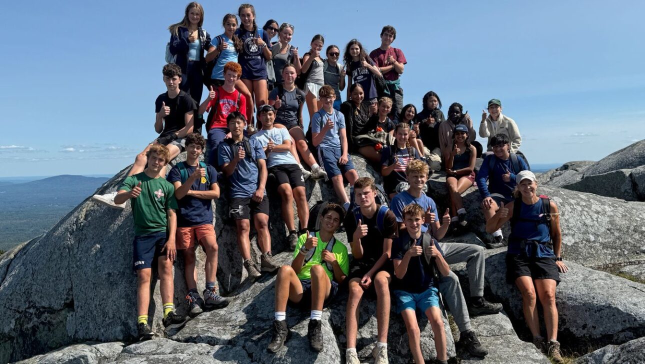 A large group of people pose together on a rocky mountaintop, wearing casual outdoor clothing and hiking gear under a clear sky.