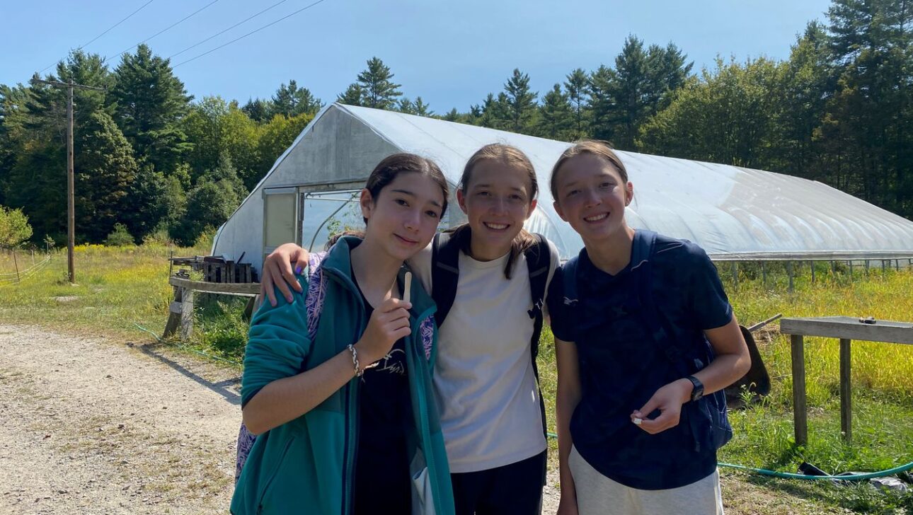 Three teenagers stand together outdoors in front of a greenhouse, surrounded by greenery and trees, smiling at the camera on a sunny day.