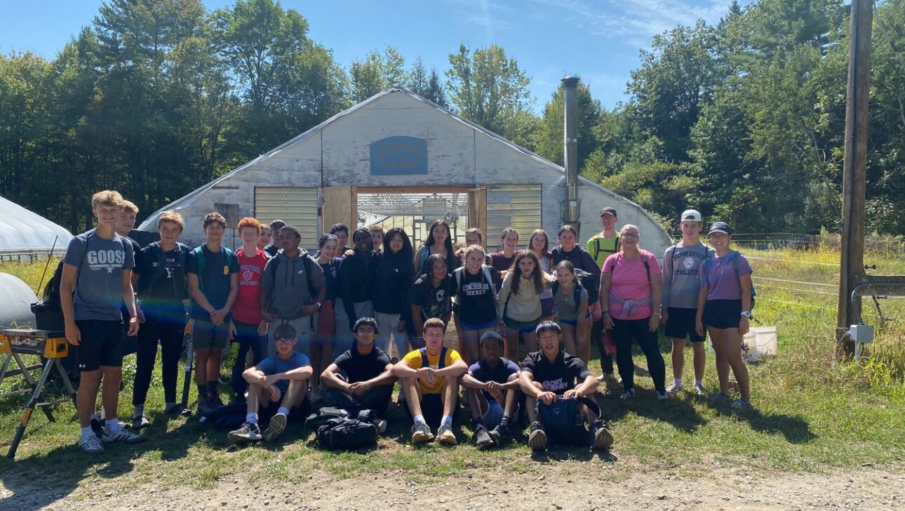 A group of young people and adults pose in front of a large greenhouse on a sunny day, with trees and blue sky in the background.