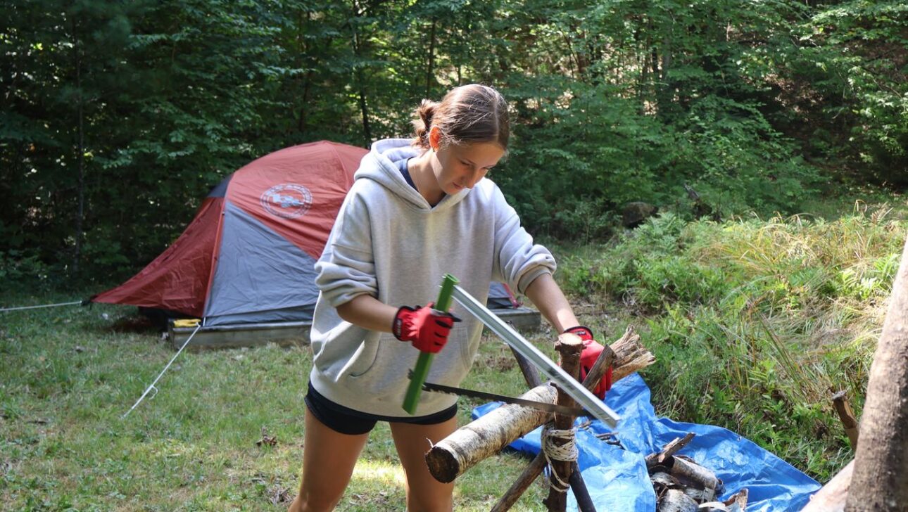 A person wearing gloves and a hoodie uses a saw to cut branches, with a tent and blue tarp behind them in a wooded outdoor setting.