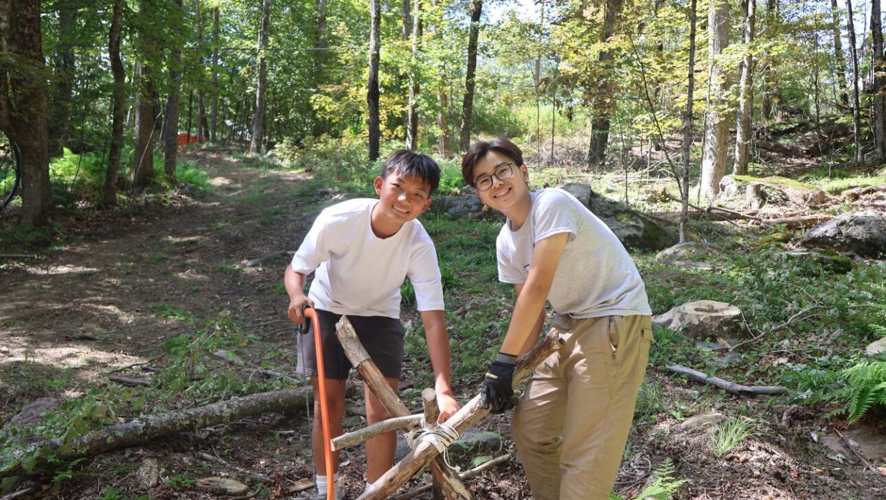 Two boys in a forest are smiling at the camera while building a wooden tripod structure with rope. Sunlight filters through the trees around them.
