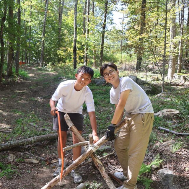 Two boys in a forest are smiling at the camera while building a wooden tripod structure with rope. Sunlight filters through the trees around them.