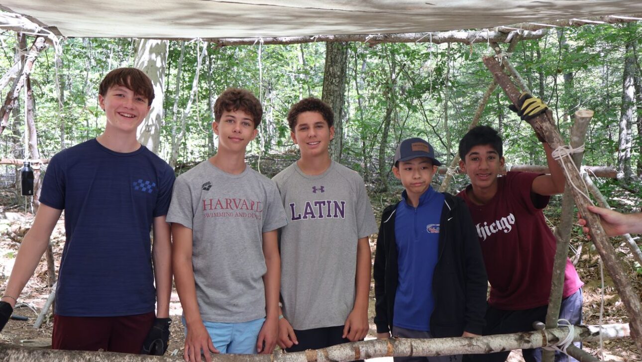 Five boys stand together under a canopy in a wooded area, posing and smiling behind a frame made of wooden branches.