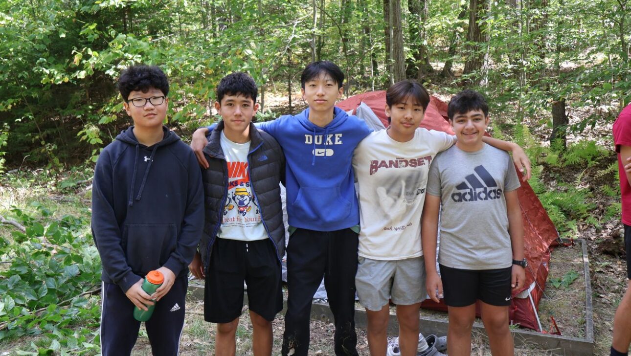 Five boys stand side by side in front of a red tent in a wooded area, dressed in casual clothes, posing for a group photo outdoors.
