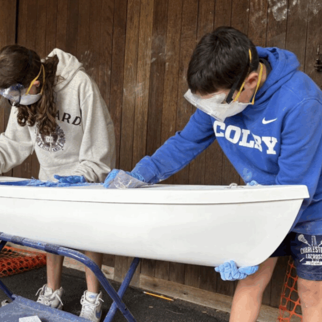 Two children wearing goggles and gloves sand a white boat hull placed on a stand, with orange safety netting and a wooden wall in the background.
