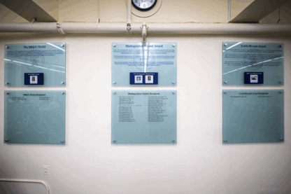 Three glass plaques mounted on a wall display the titles and lists of recipients for the IBM’s Nobel, Distinguished Alumni, and Louis Boyars Awards beneath a clock.
