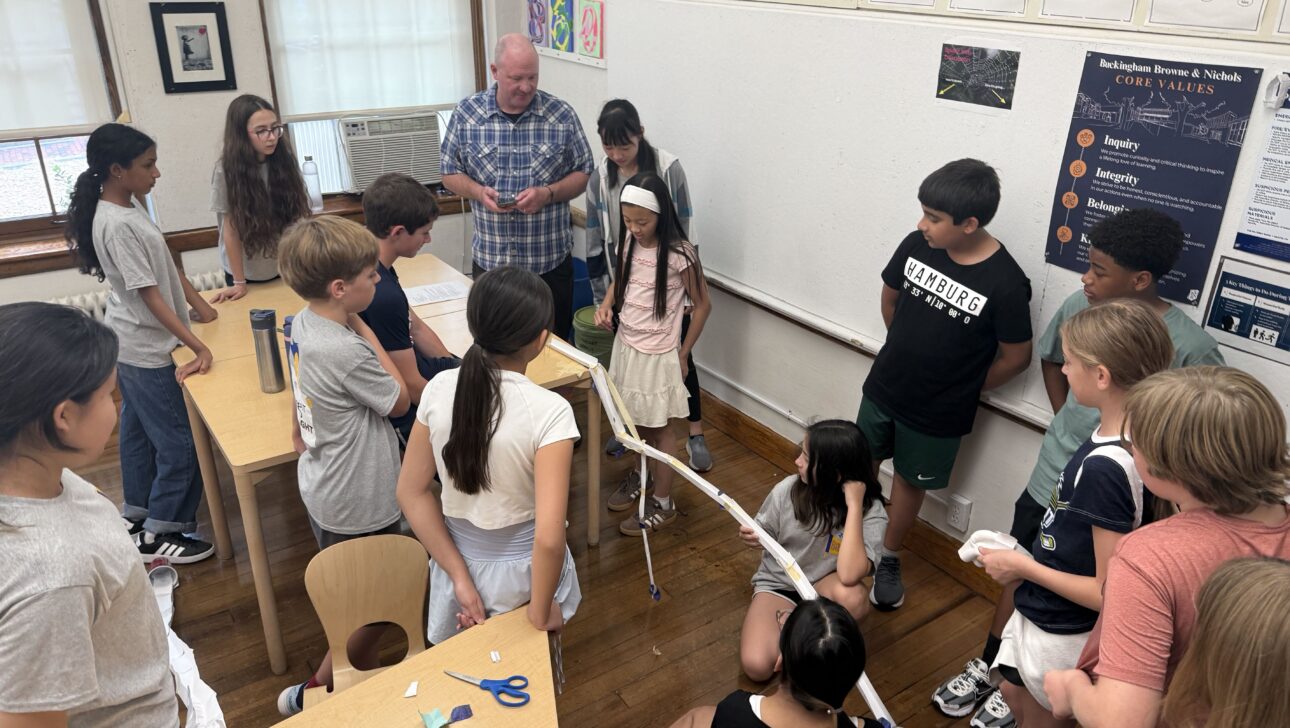 A group of students and a teacher gather around a classroom project involving a long paper structure laid out across the desks and floor.