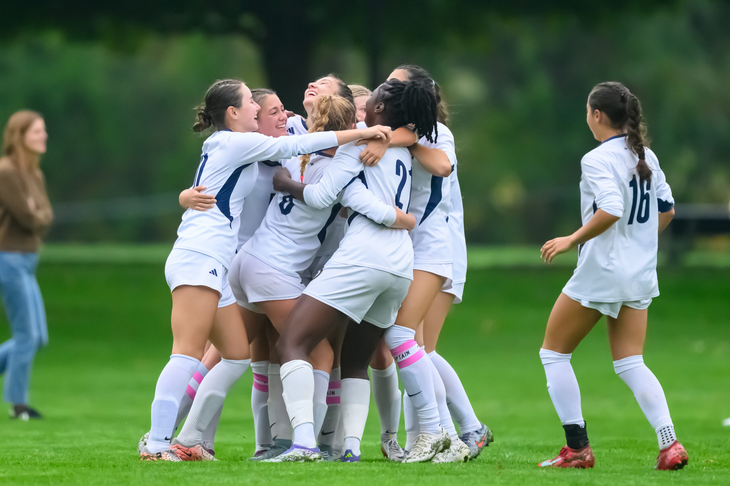 A group of female soccer players in white uniforms huddle and celebrate on a green field, while another player and a person in casual clothes stand nearby.