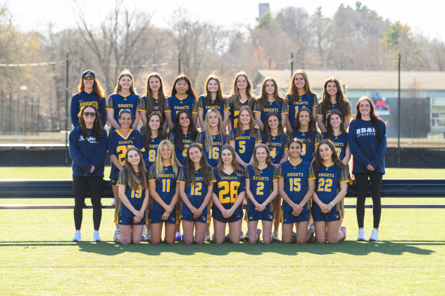A women’s lacrosse team poses outdoors in three rows on a turf field, wearing matching blue and yellow uniforms, with two coaches standing at each end.