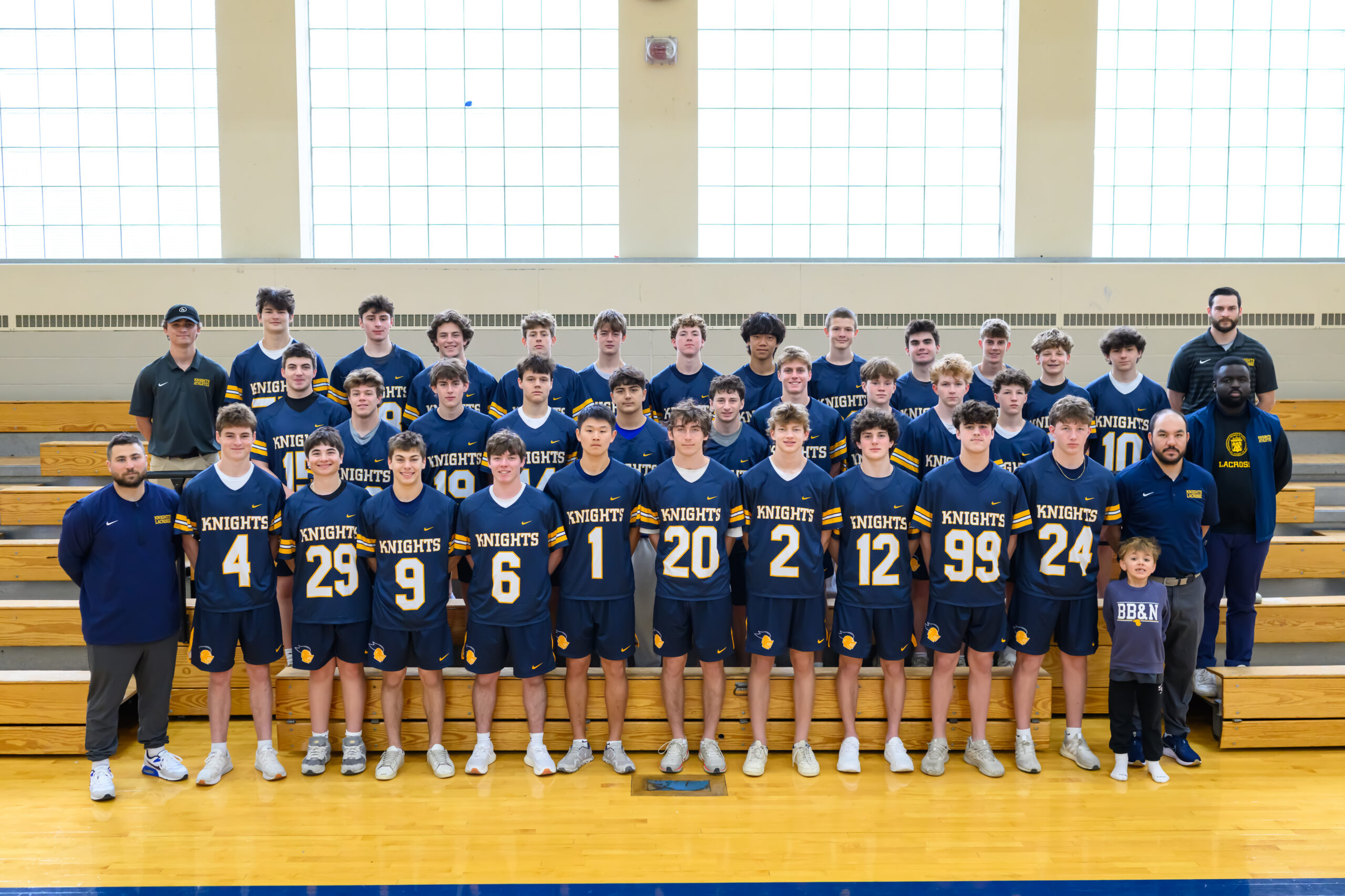 A boys’ lacrosse team poses for a group photo in uniform on bleachers inside a gym, with coaches and a young child standing with them.