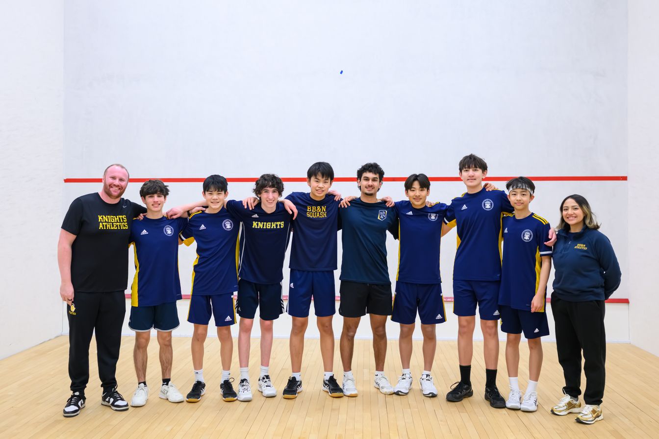 Nine students and two adults in athletic attire stand together on a squash court, posing for a group photo in front of a white wall with red boundary lines.
