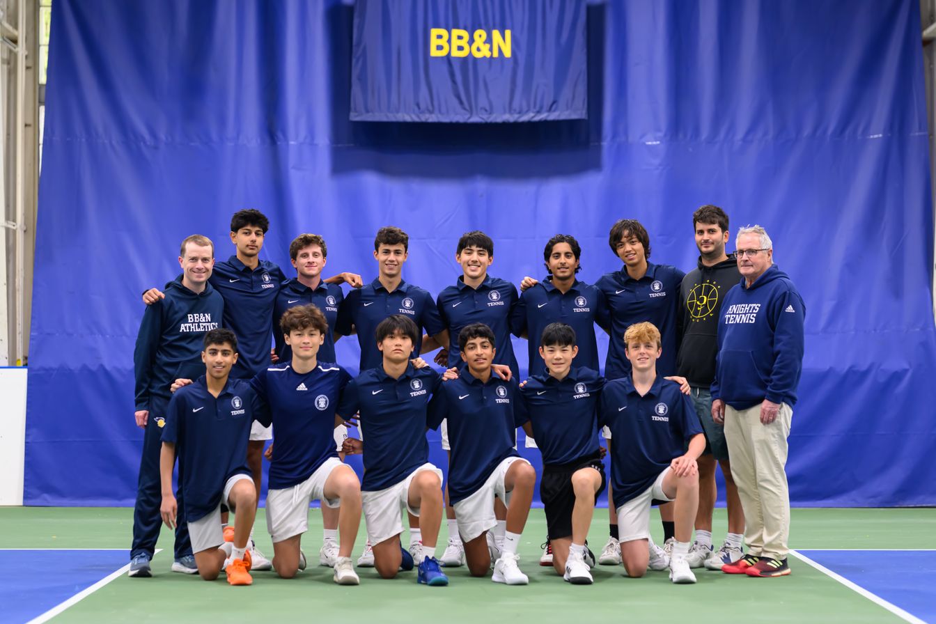 A group of thirteen boys in matching navy tennis uniforms and two adults pose together on an indoor tennis court under a BB&N banner.