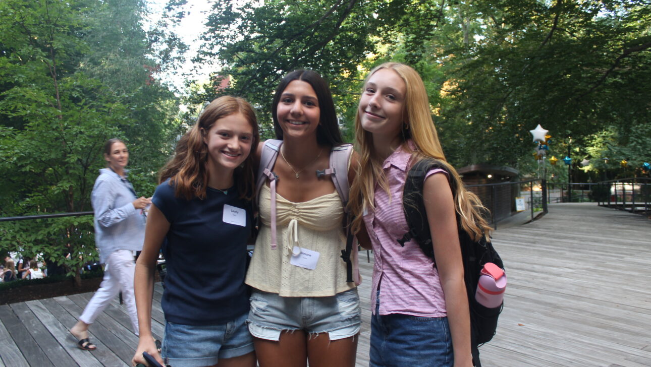 Three teenage girls with backpacks and name tags stand together on a wooden walkway outdoors, smiling at the camera. Trees and another person are in the background.