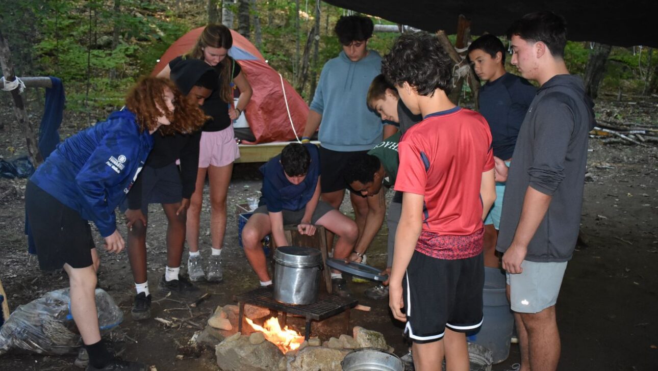 A group of teenagers stand around a campfire under a tarp, cooking with metal pots and grilling food, with a tent and trees in the background.
