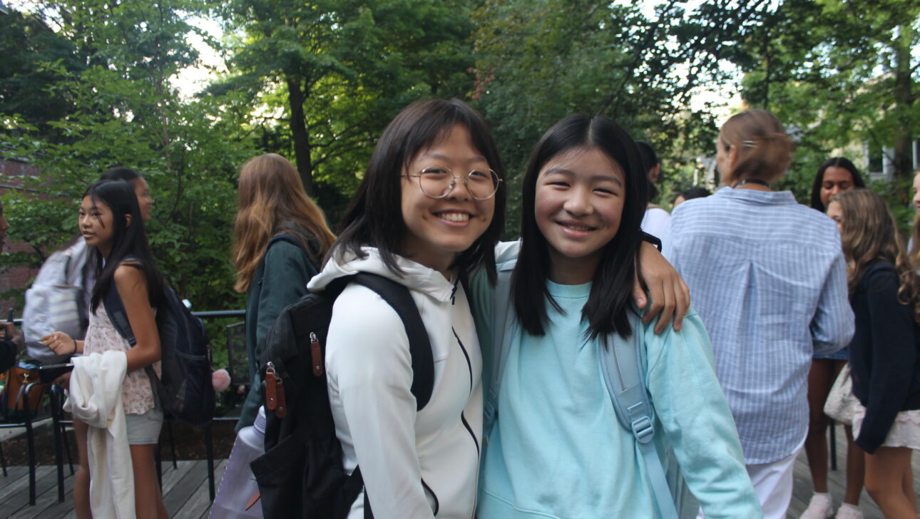 Two girls with backpacks smile and pose together outdoors, surrounded by several other people in casual clothing with a background of trees.