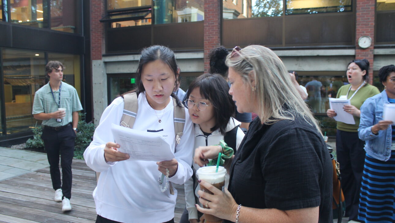 Three people stand outside, closely examining a sheet of paper. Others with papers are visible in the background near a building with large windows.