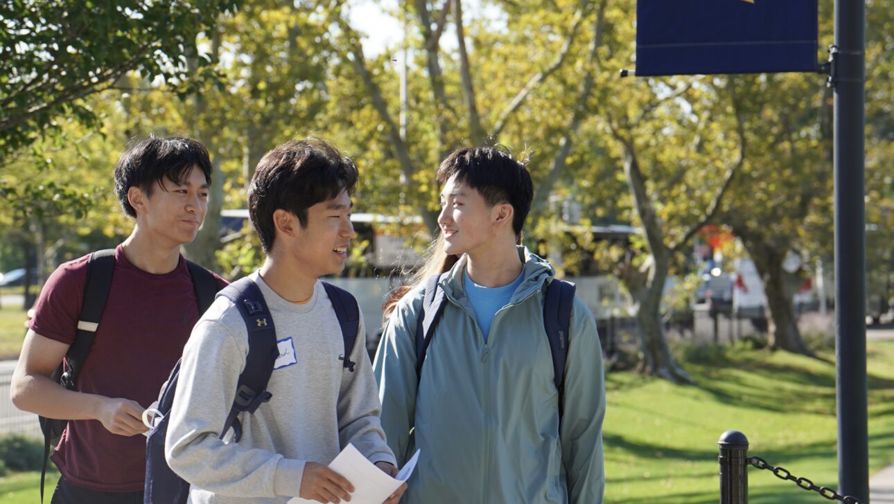 Three students with backpacks walk outside on a sunny day near a campus banner featuring a knight helmet logo.