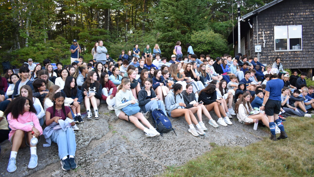 A large group of teenagers sit outdoors on stone steps, some talking or looking at their phones, surrounded by trees and a wooden building in the background.