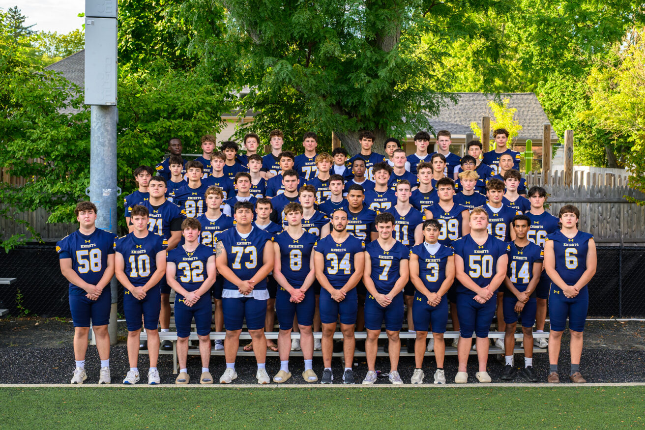 A high school football team poses outdoors in blue and gold uniforms, standing in rows with trees and a wooden fence in the background.
