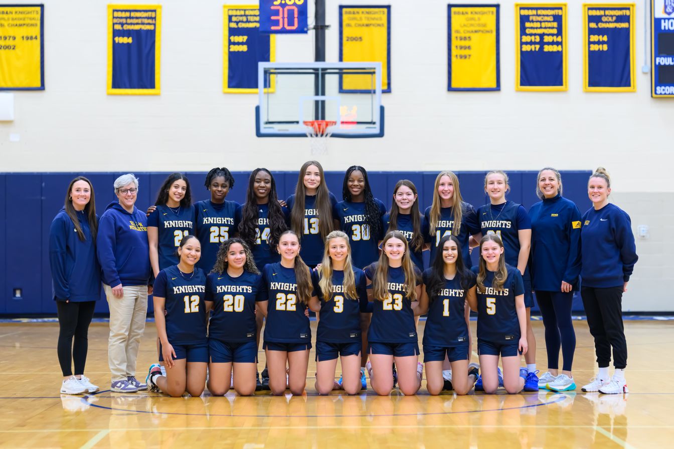 A girls’ volleyball team and coaches pose in uniform on an indoor court, with banners and a basketball hoop visible in the background.