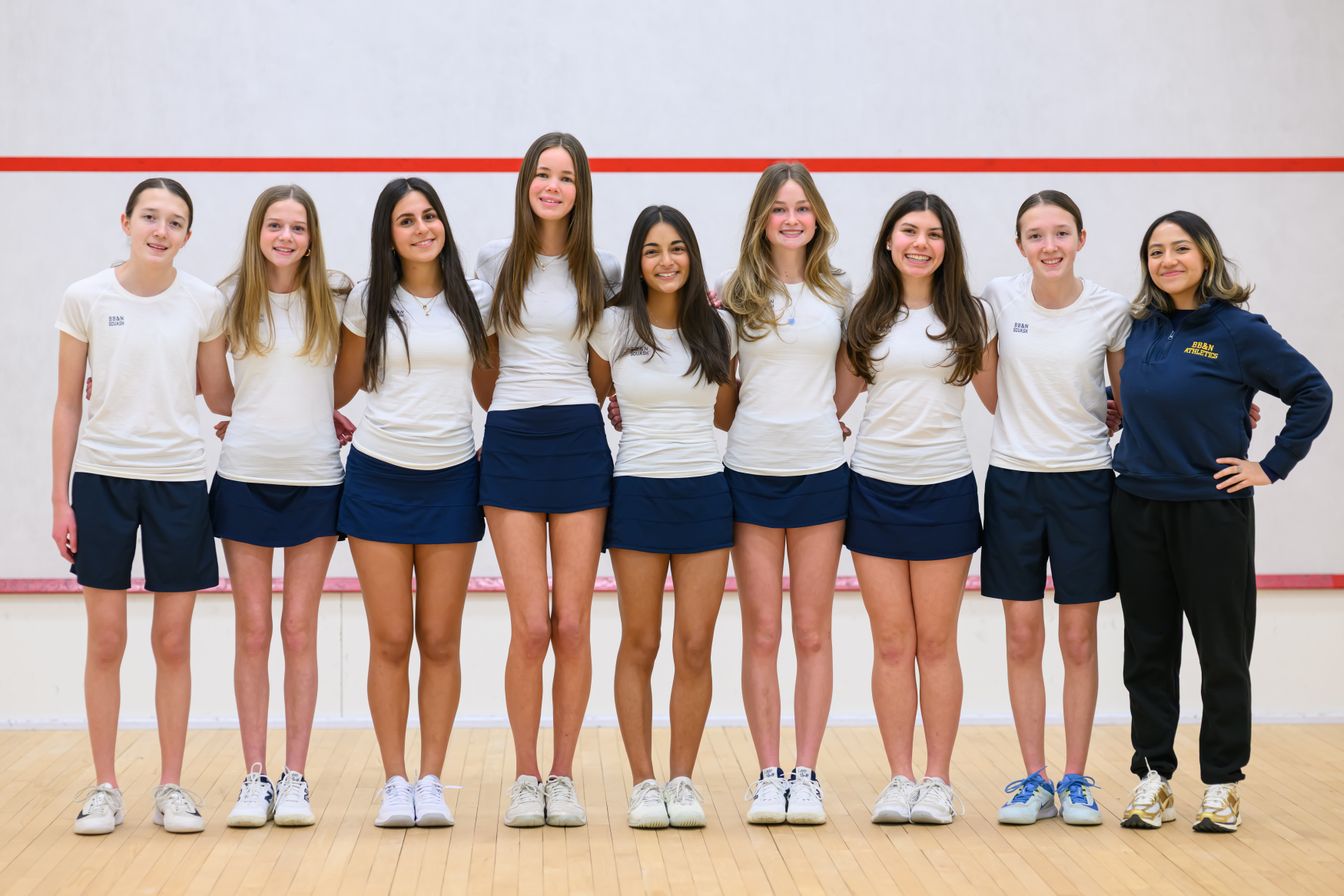 Eight young women in white shirts and navy skirts stand in a row on a squash court, with one coach in dark athletic wear standing beside them.