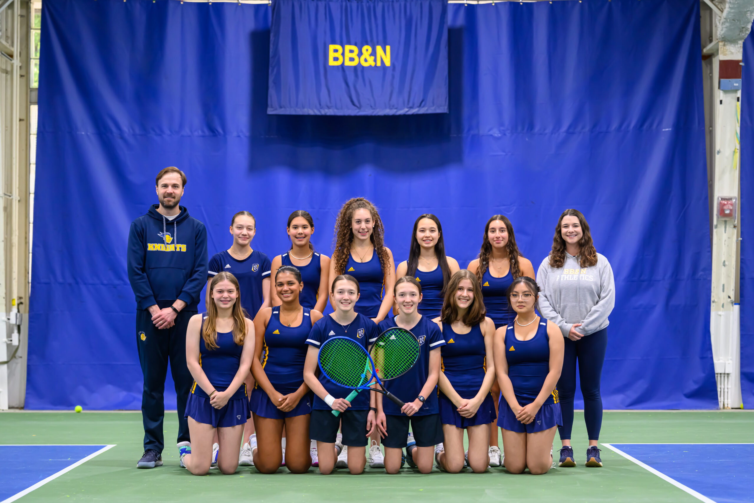 A girls’ tennis team poses indoors with their coach and another adult in front of a blue curtain with a “BB&N” sign. Two players in front hold green tennis rackets.