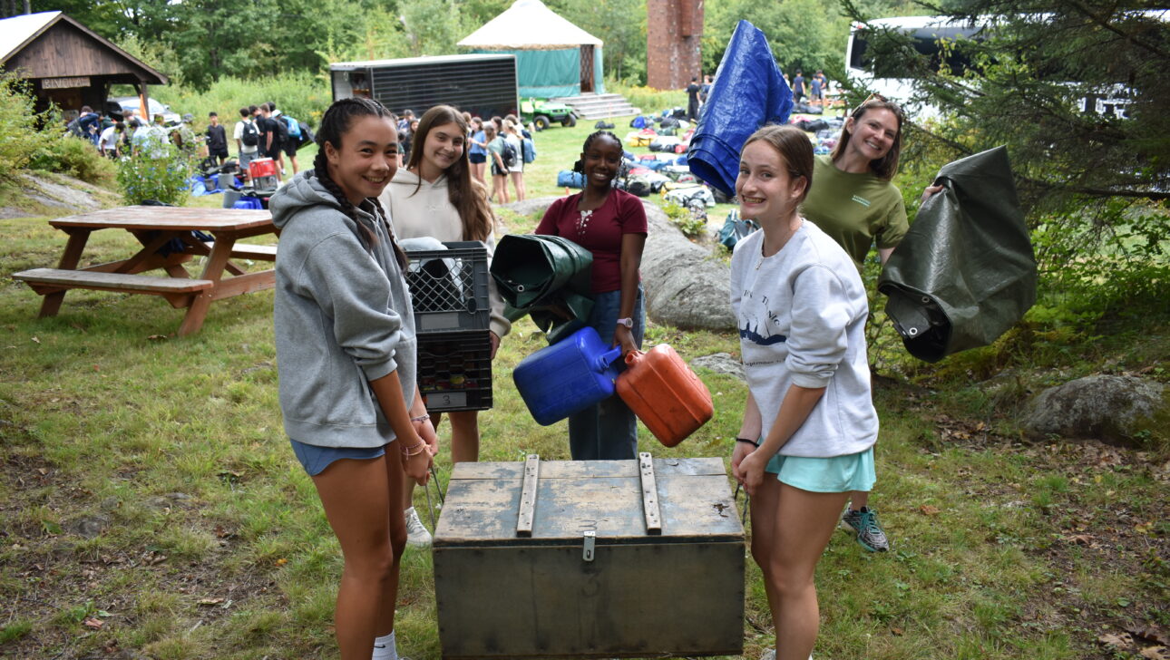 Five young people outdoors carry camping gear and supplies, including a large chest, gas cans, and tarps, with tents and more equipment visible in the background.