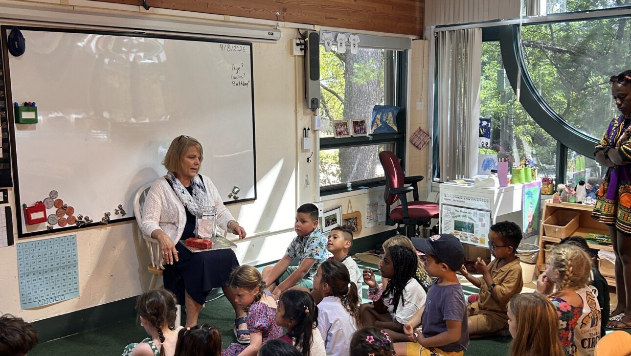 A teacher sits in front of a group of young children on the floor in a bright classroom with large windows and wooden ceiling.
