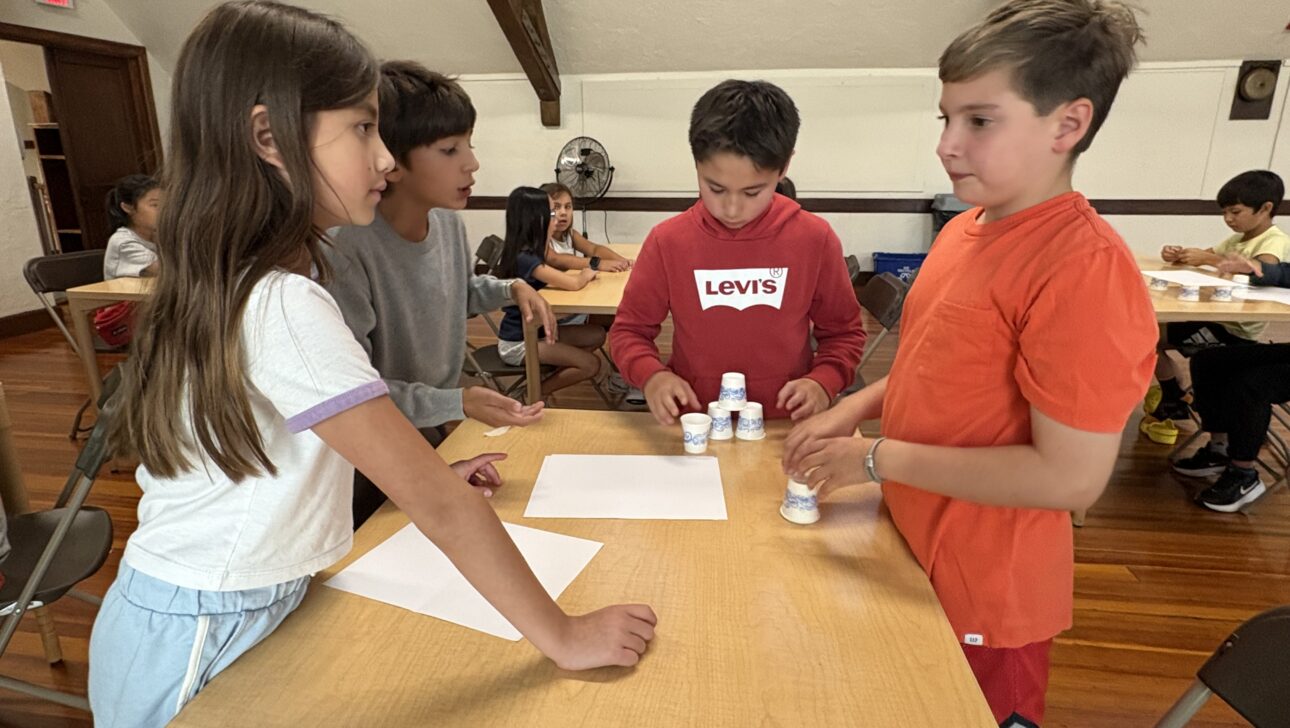 Four children stand around a table with paper and stacked plastic cups, engaging in an activity in a room with other children in the background.