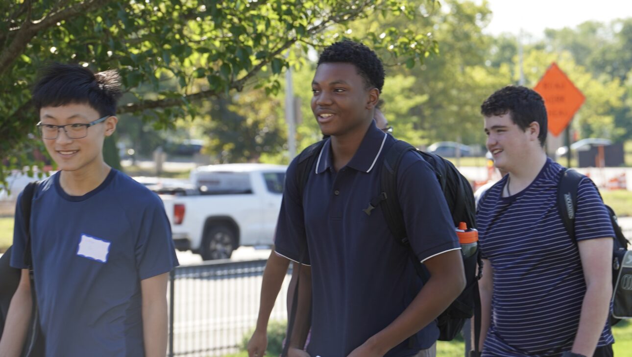 Three students walk outside on a sunny day, carrying papers and backpacks, with trees and a white pickup truck visible in the background.
