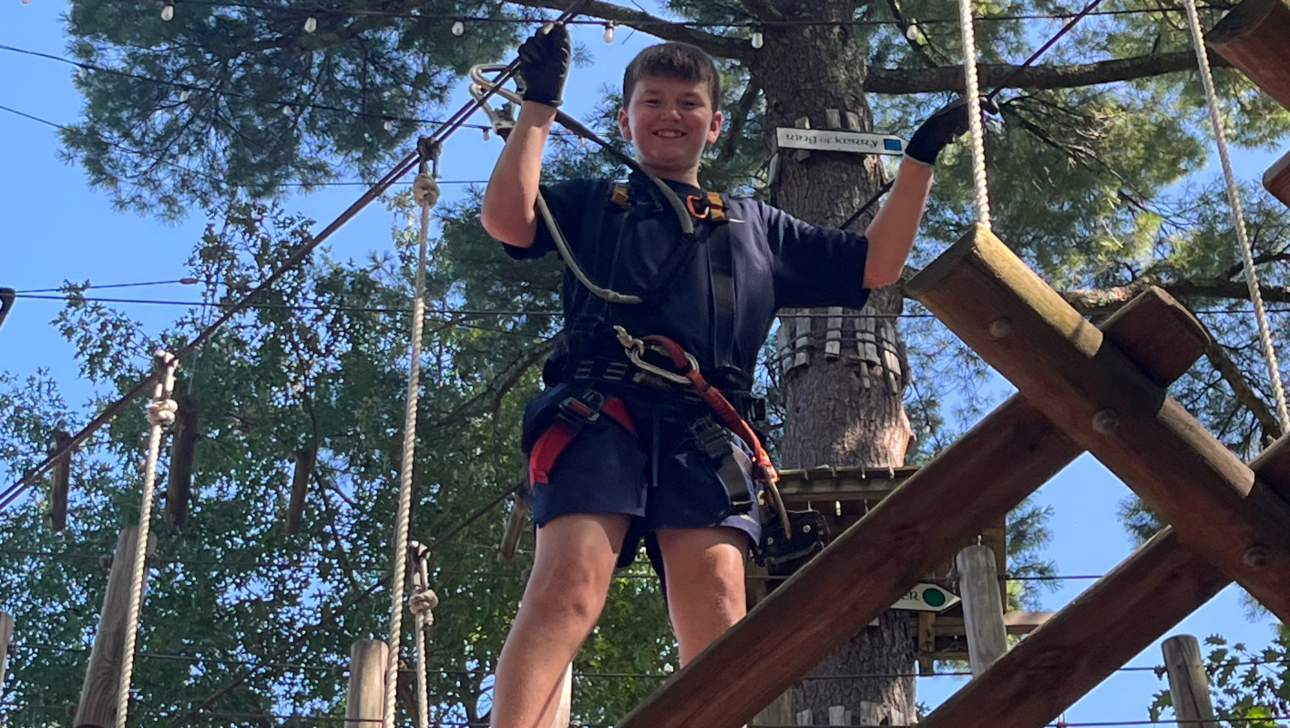 A boy wearing a harness and gloves stands on a wooden platform during a ropes course, surrounded by trees and blue sky.